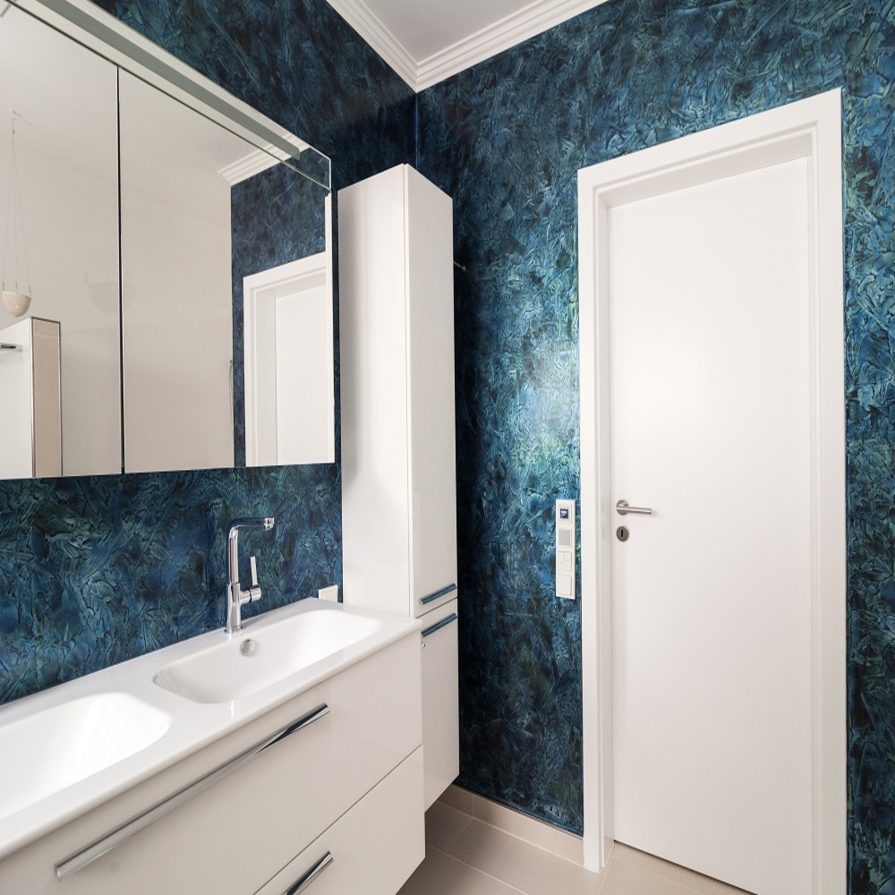 Ein modernes Badezimmer mit blau getöntem Marmor-Wandmuster, das einen edlen Touch verleiht. Auf der linken Seite befindet sich ein Waschtisch mit einem großen Spiegel darüber. Neben dem Waschtisch gibt es einen hohen weißen Schrank, der zusätzlichen Stauraum bietet. Gegenüber dem Waschtisch befindet sich eine weiße Tür. Die Kombination von Weiß und dem blauen Muster gibt dem Raum eine erfrischende und gleichzeitig luxuriöse Atmosphäre. Das Design wirkt schlicht, aber dennoch stilvoll und modern.